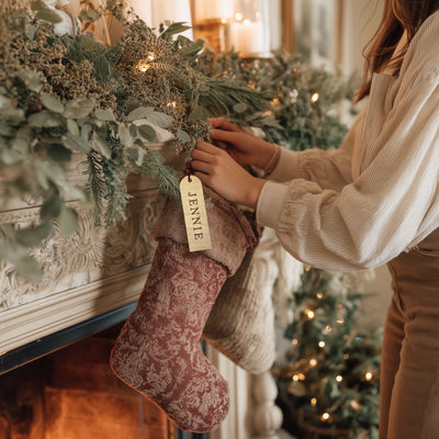 Person placing a Christmas stocking labeled 'Jennie' on a fireplace mantle with a decorated tree in the background.