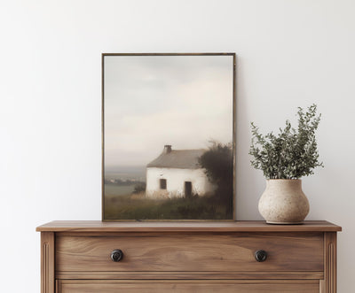 Framed artwork of a cottage on a wooden dresser with a vase of greenery.